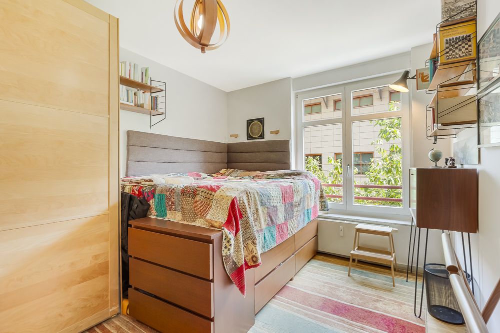 Helles Schlafzimmer mit hohem Bett, bunter Decke, Kommode und großem Fenster.