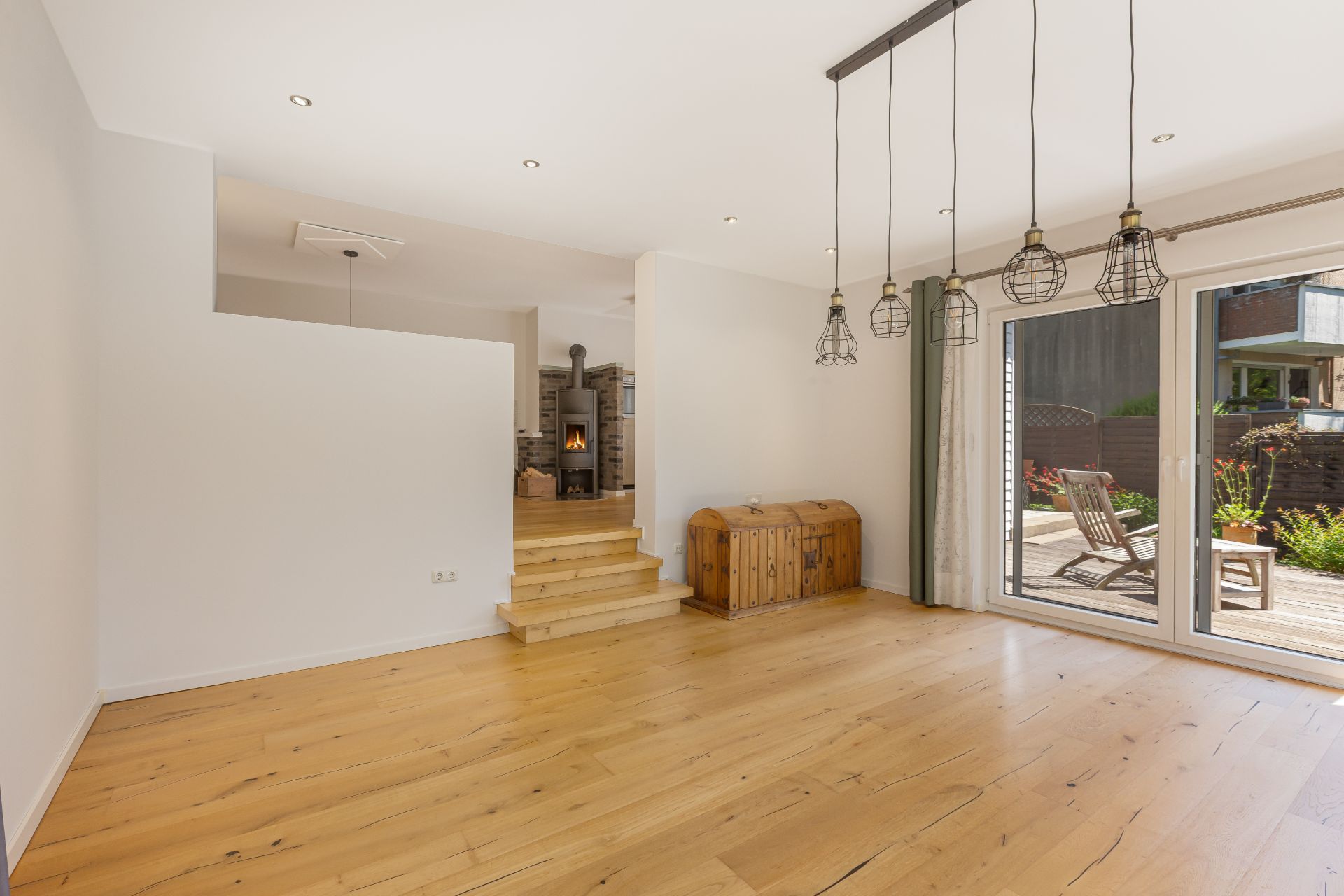 Heller Wohnraum mit Holzboden, Hängelampen, Holztruhe und Zugang zur Terrasse mit Gartenmöbeln.