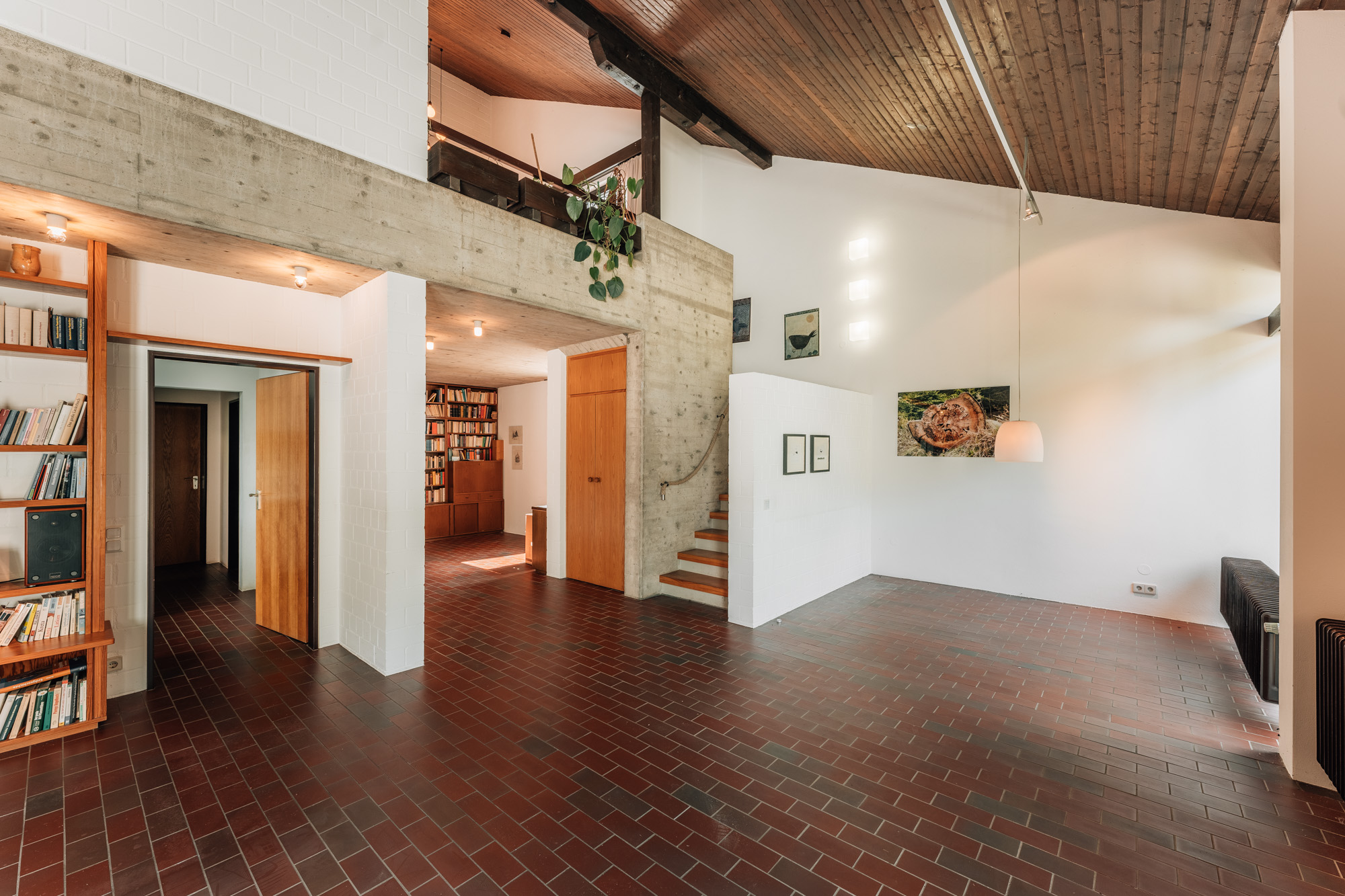 Wohnzimmer mit roten Fliesen, Holzdecke, Treppe, Bücherregalen und Wandbildern.