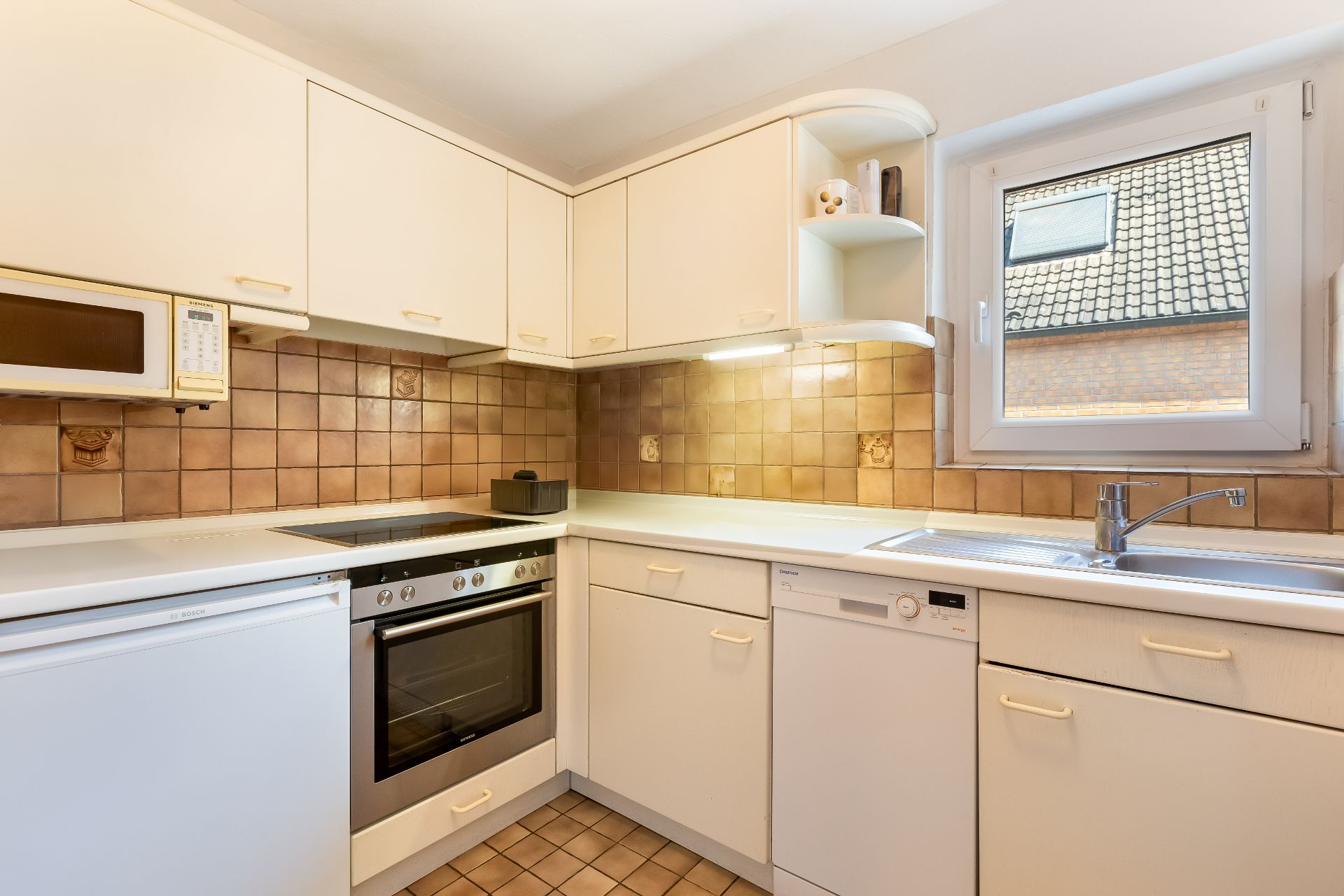 Helle Küche mit weißen Schränken, Herd, Backofen, Spüle, Geschirrspüler und Fenster.