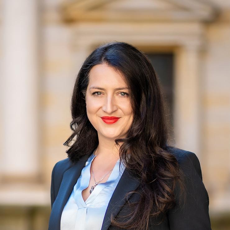 Frau mit langem dunklem Haar im Anzug steht draußen vor unscharfem Gebäude.