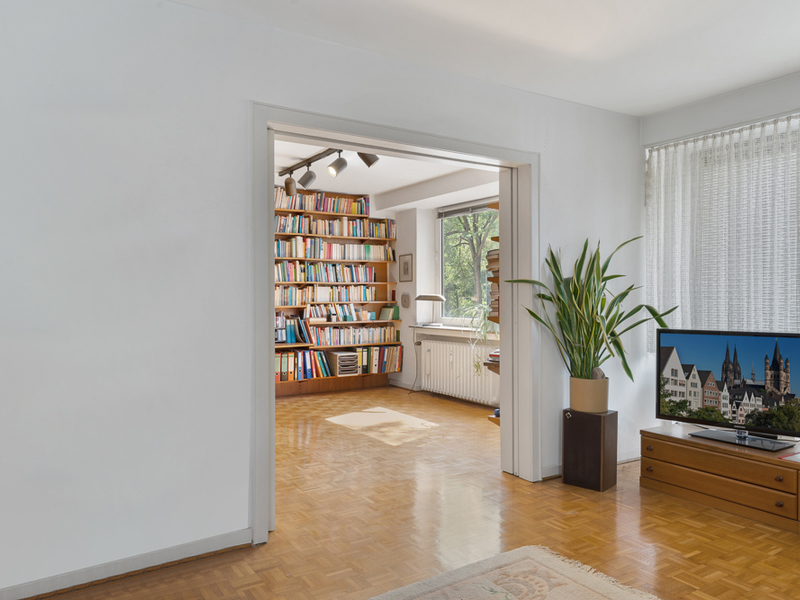 Wohnzimmer mit Fernseher, Zimmerpflanze und Blick auf Regal mit vielen Büchern im Nebenraum.
