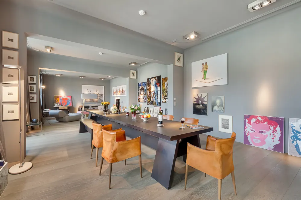 Modernes Esszimmer mit großem Tisch, sechs Stühlen und bunter Kunst an grauen Wänden.