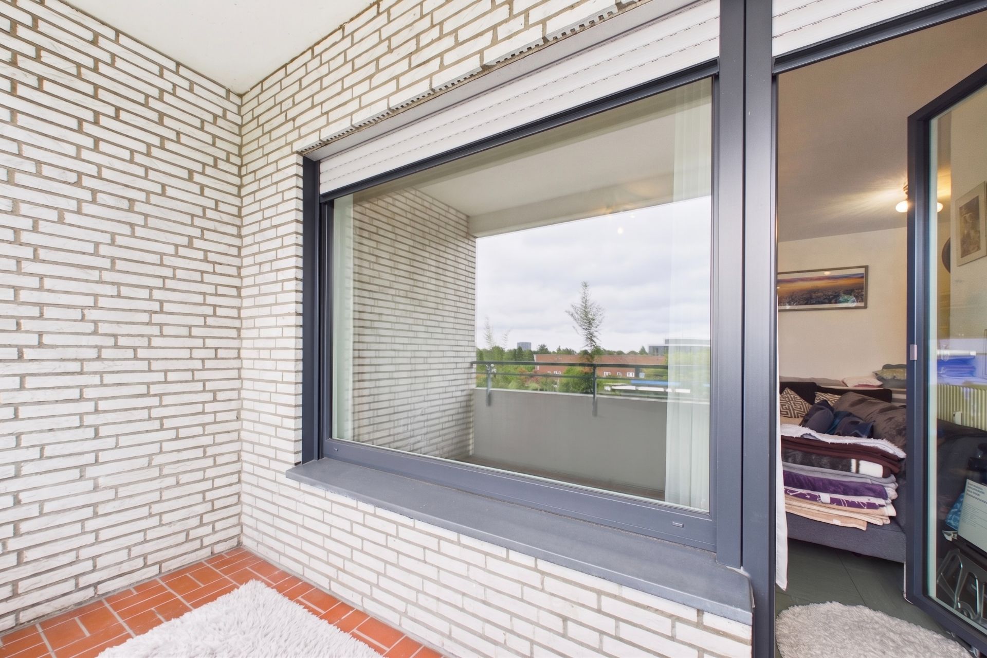 Balkon mit weißen Klinkerwänden, großem Fenster und Blick in ein Schlafzimmer.