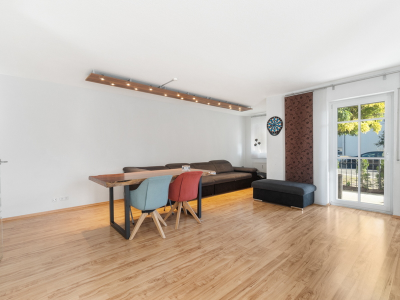 Wohnzimmer mit Holzboden, Esstisch, bunten Stühlen, Sofa und großer Fensterfront.