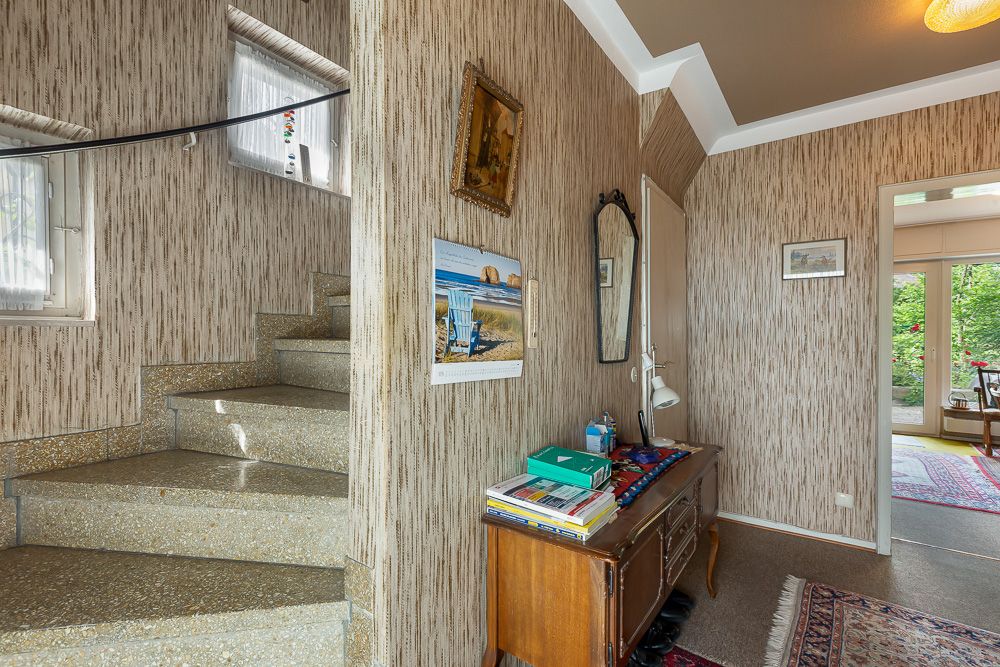 Hausflur mit Treppe, Kommode, Wandspiegel und Blick in ein helles Wohnzimmer.