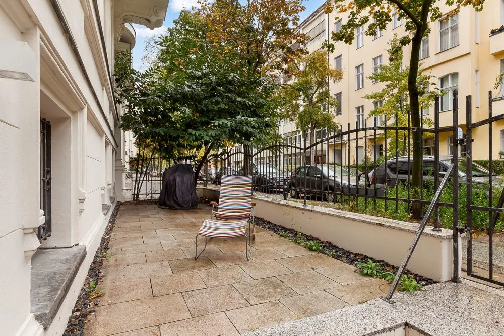 Gepflasterte Terrasse mit einem Gartenstuhl, Baum und schwarzem Grill, umgeben von Zaun und Pflanzen.