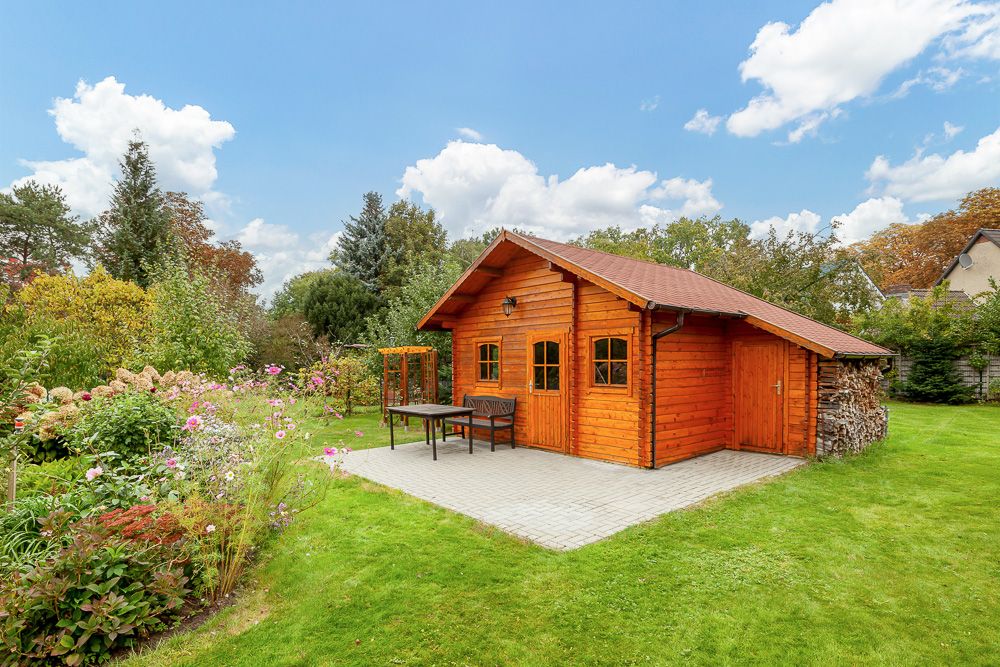 Holzgartenhaus mit Terrasse, Tisch und Bank auf einer Rasenfläche mit Blumen und Bäumen.