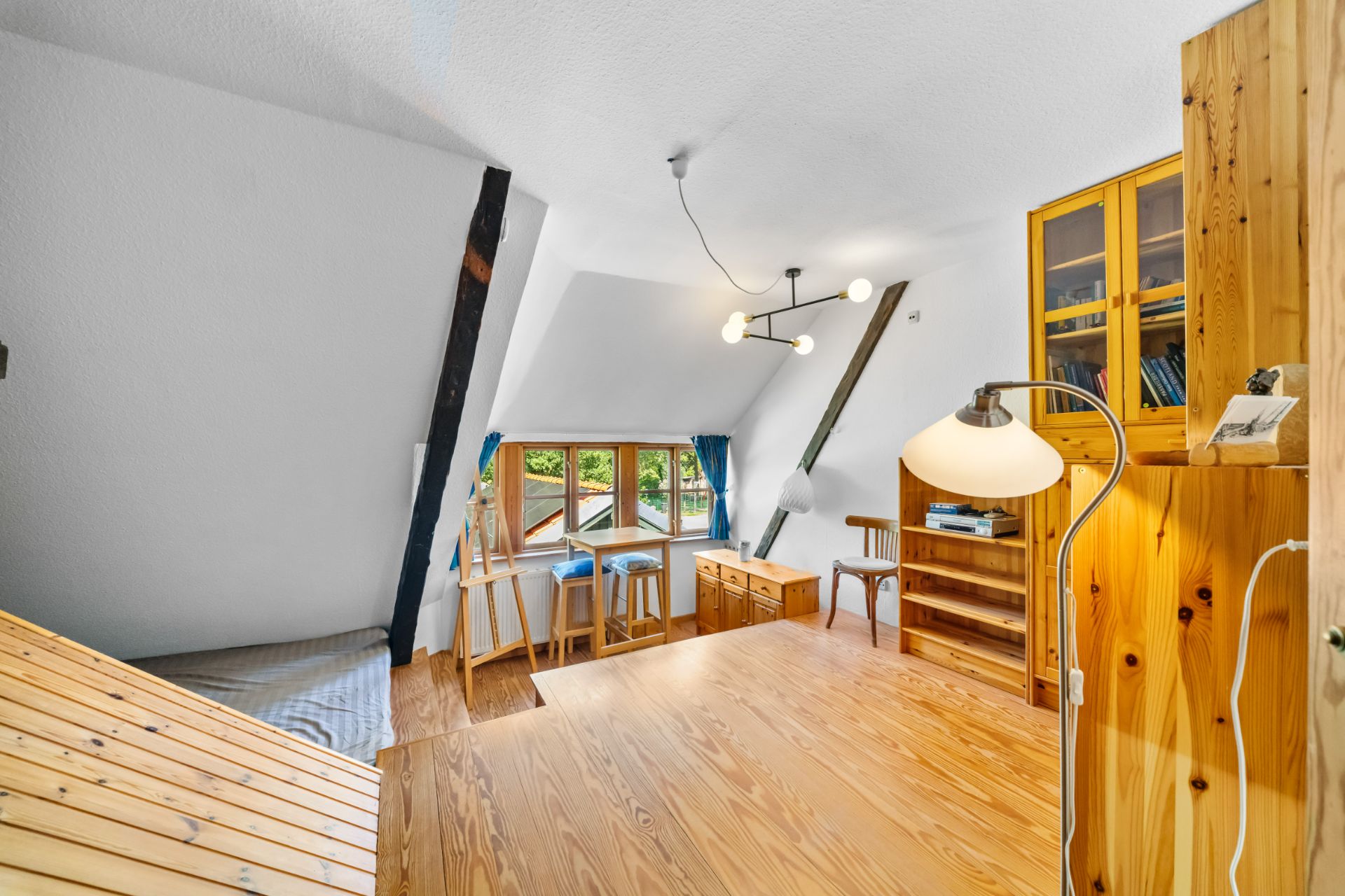 Helles Dachzimmer mit Holzfußboden, Schreibtisch am Fenster und Bücherregal.