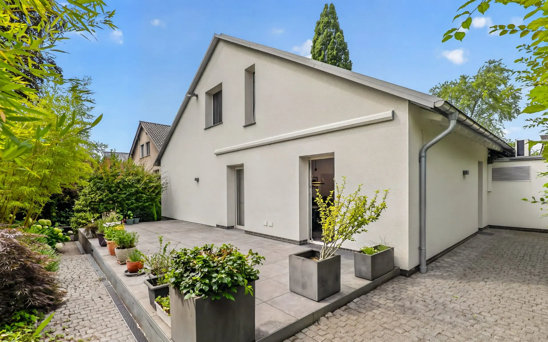 Moderne Terrasse mit Pflanzkübeln neben einem weißen Wohnhaus und gepflastertem Weg.