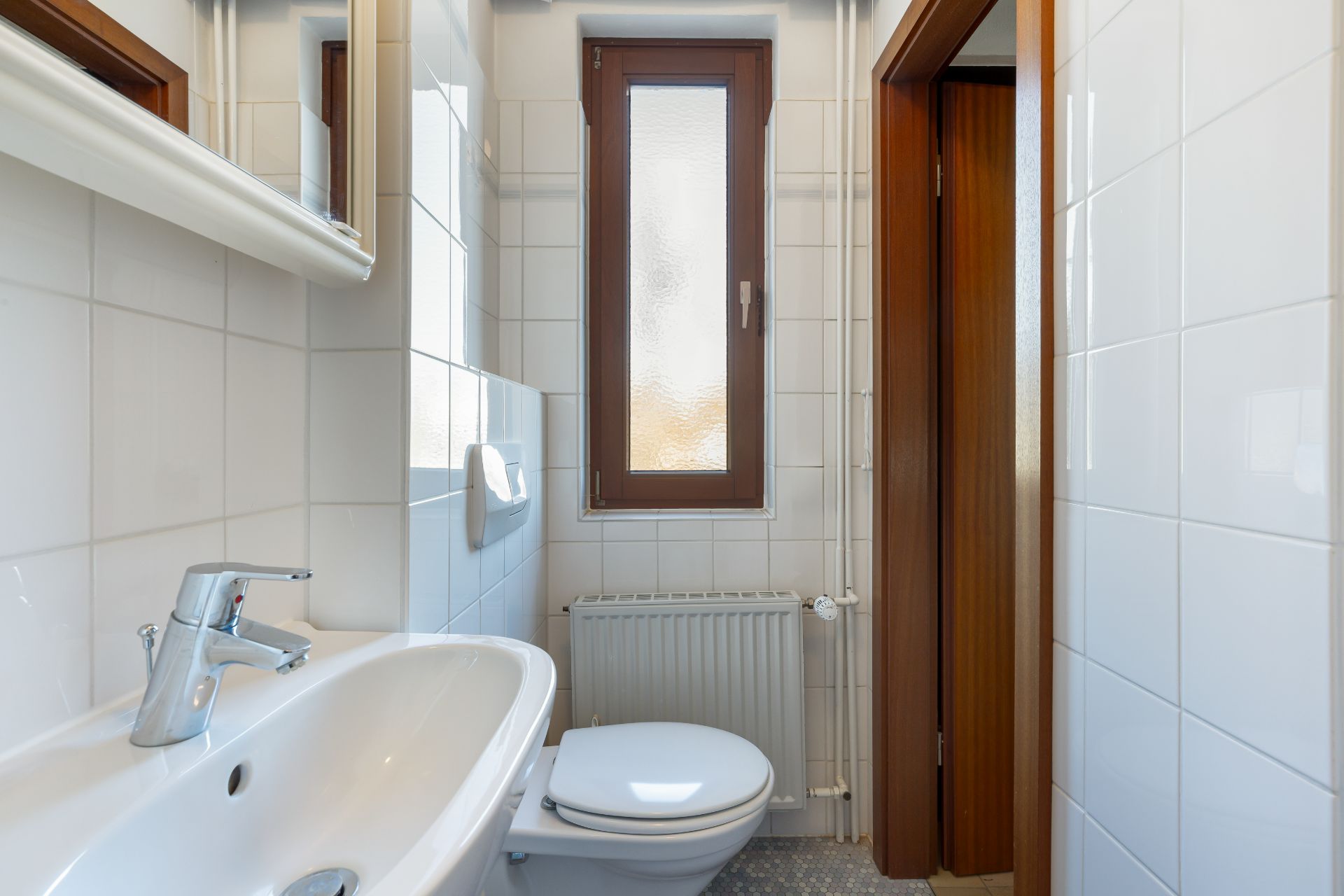 Kleines Badezimmer mit Waschbecken, Toilette und Fenster, weiße Fliesen an den Wänden.