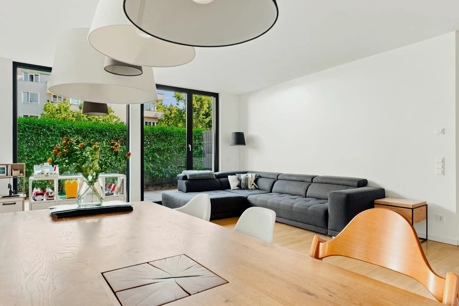 Modernes Wohnzimmer mit großer Ecksofa, Esstisch, Lampen und bodentiefen Fenstern zum Garten.