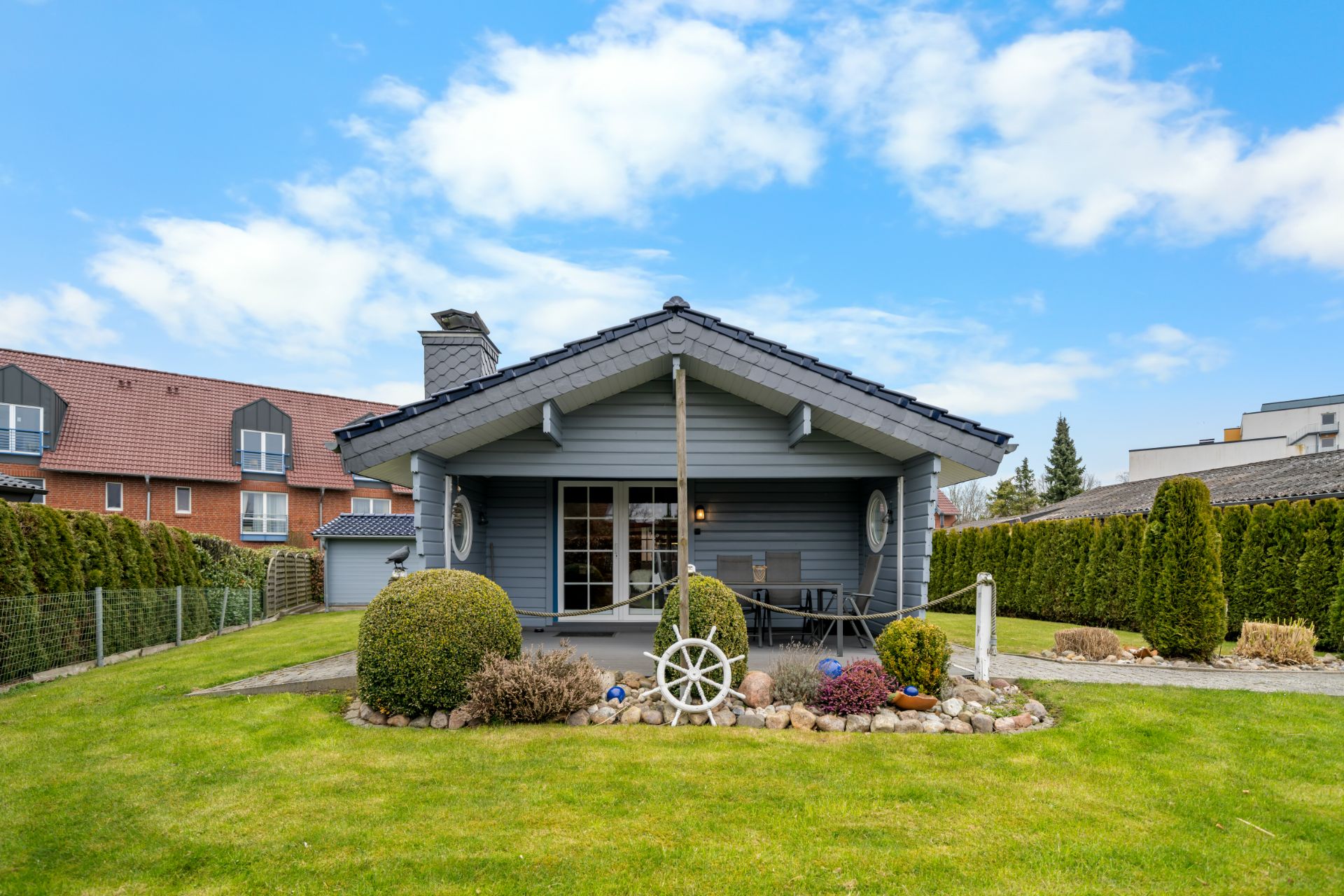 Ein einstöckiges graues Haus mit Terrasse und Garten, umgeben von Sträuchern und Rasen.