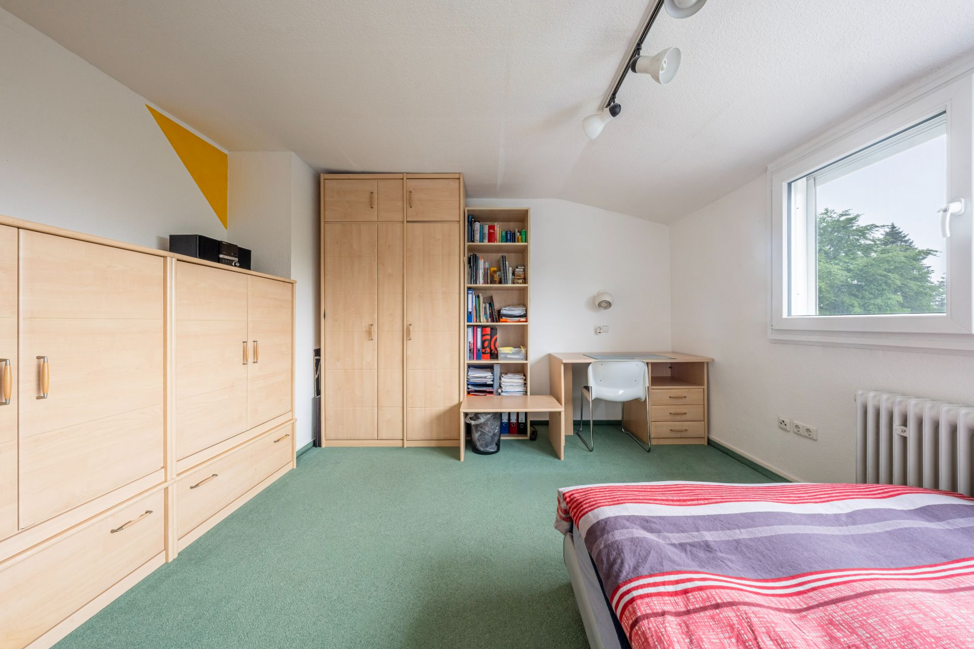 Schlafzimmer mit Holzmöbeln, grünem Teppich, Schreibtisch und Fenster mit Blick ins Freie.