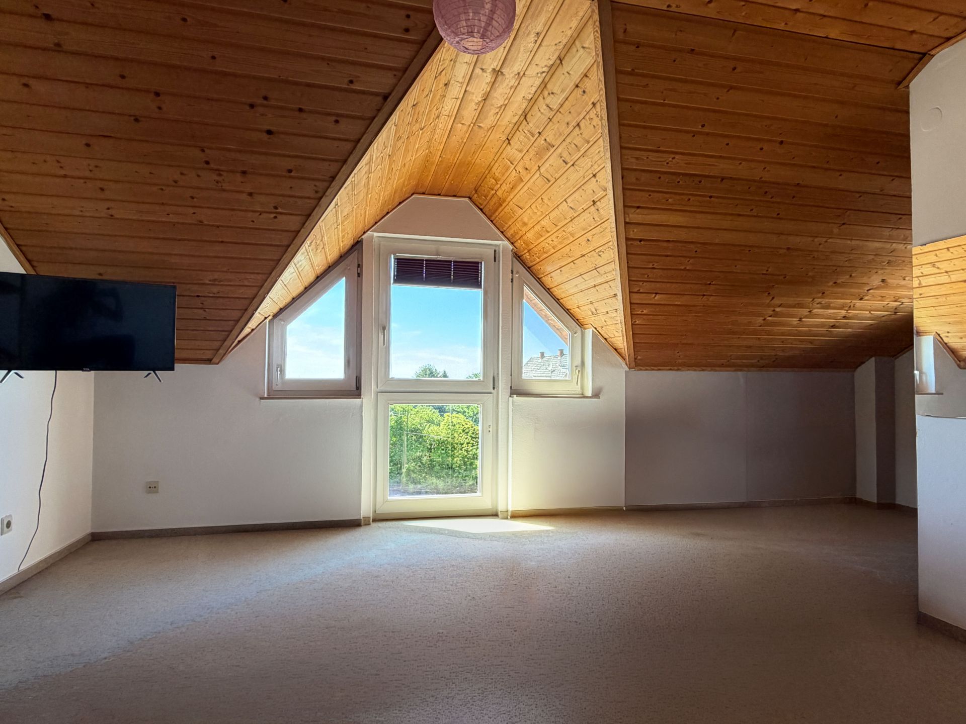 Helles Dachzimmer mit Holzdecke, großen Fenstern und einem Fernseher an der Wand.
