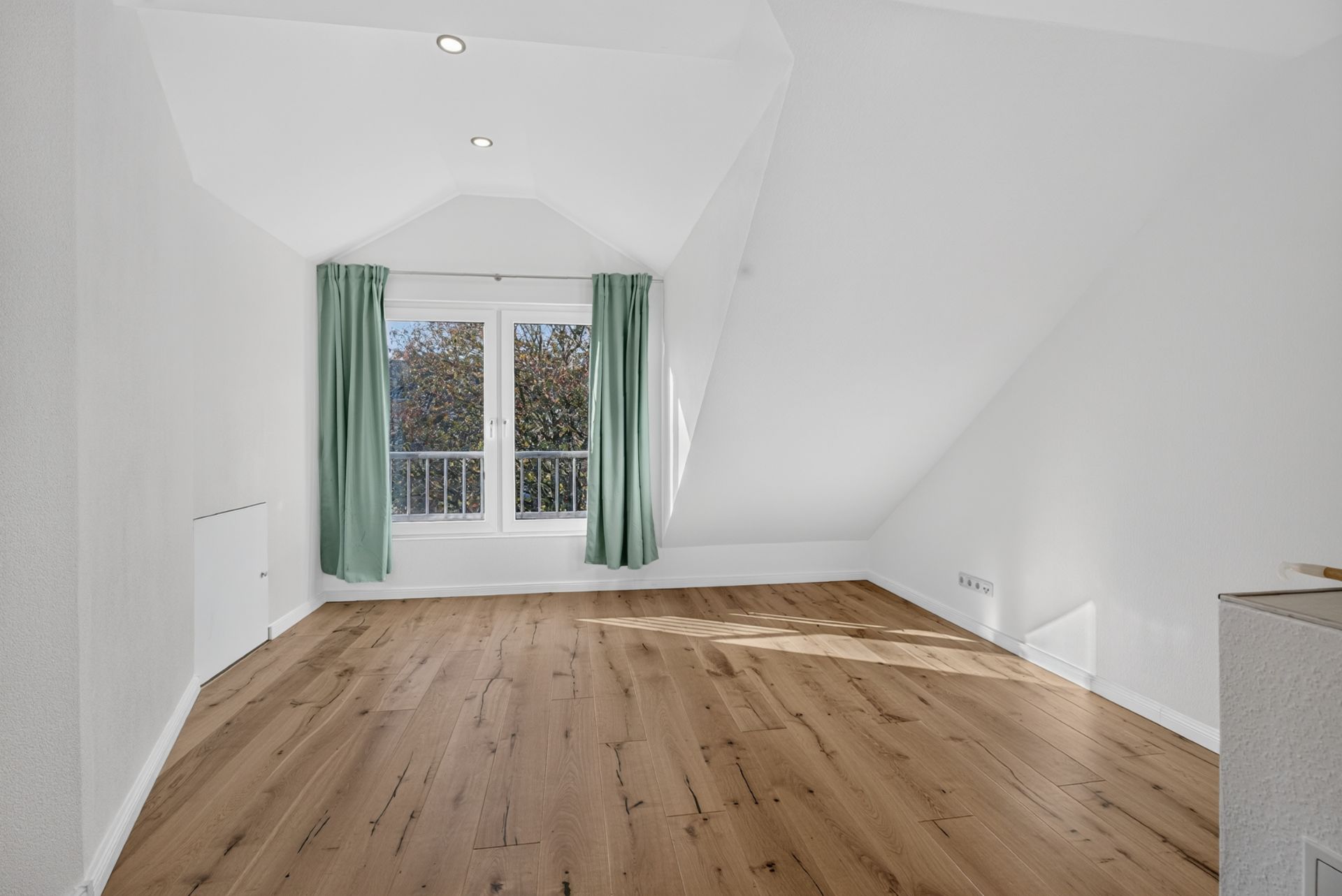 Helles Zimmer mit Holzfußboden, Dachschrägen und Fenster mit grünen Vorhängen.