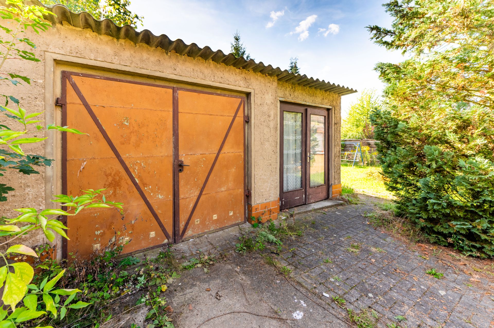 Garage mit orangefarbenem Tor und Glastür neben Pflasterweg und Sträuchern im Garten.