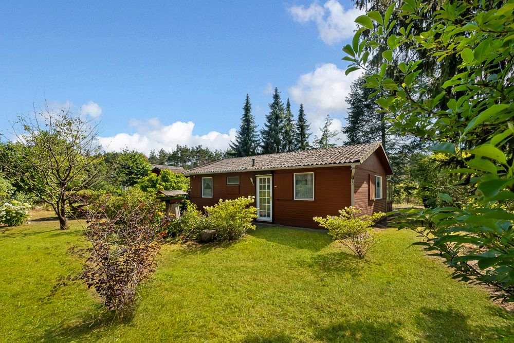 Kleines braunes Haus mit Garten, umgeben von Bäumen und Sträuchern, unter blauem Himmel.