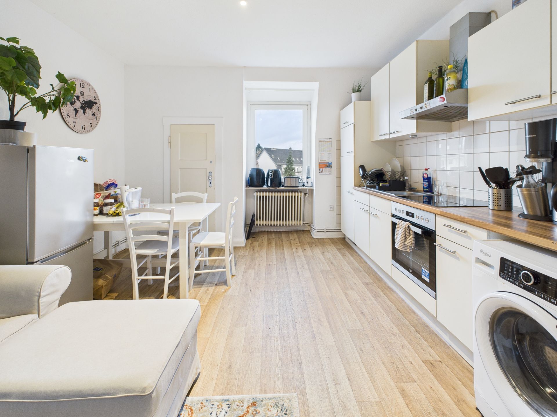 Helle Küche mit weißen Schränken, Esstisch, Sofa und großem Fenster.