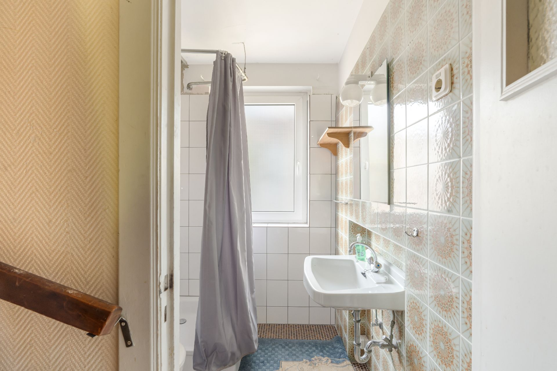 Kleines Badezimmer mit Dusche, Waschbecken, Spiegel und gemusterter Fliesenwand.