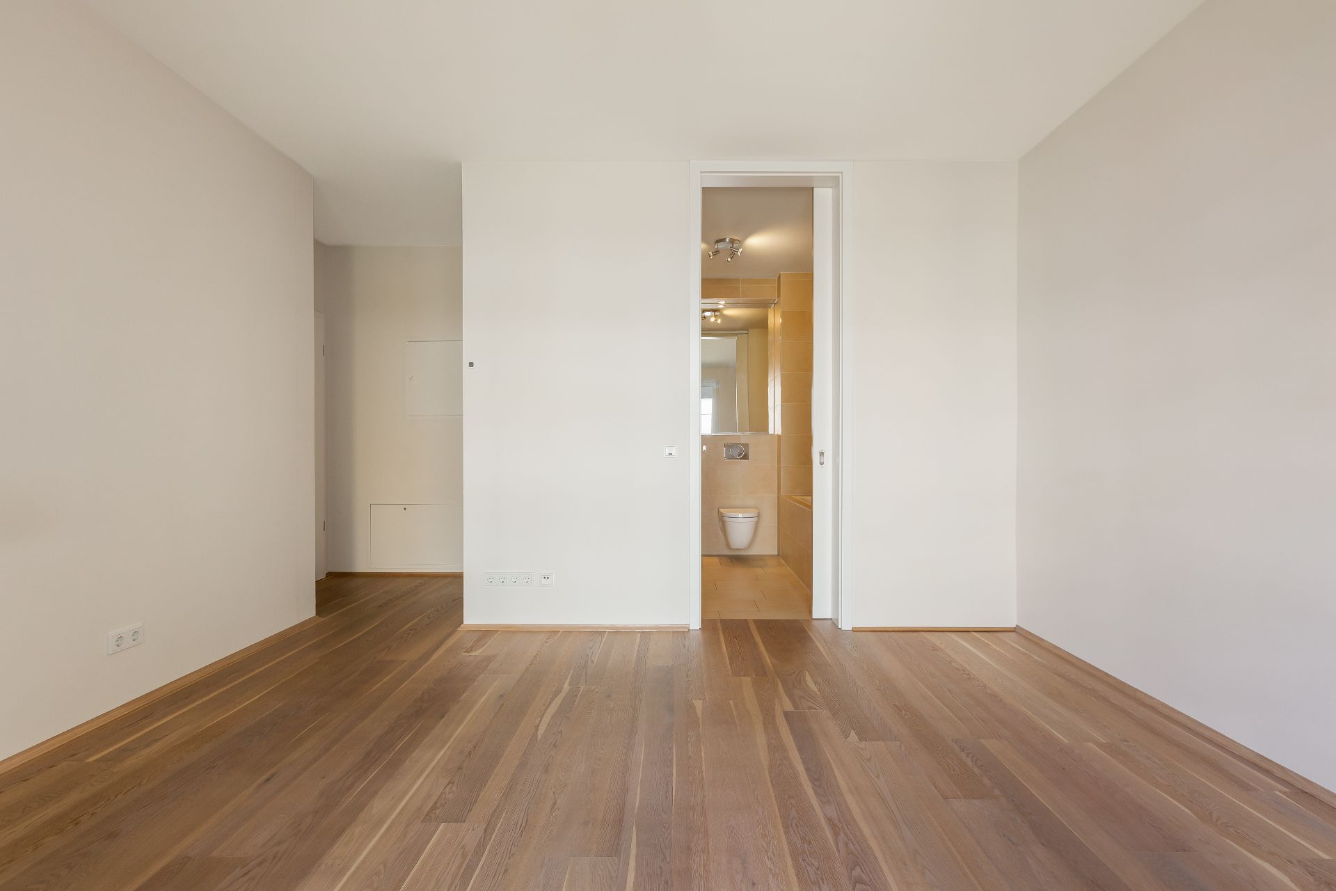 Leerer Raum mit Holzboden und Blick auf ein Badezimmer mit WC und Spiegel.