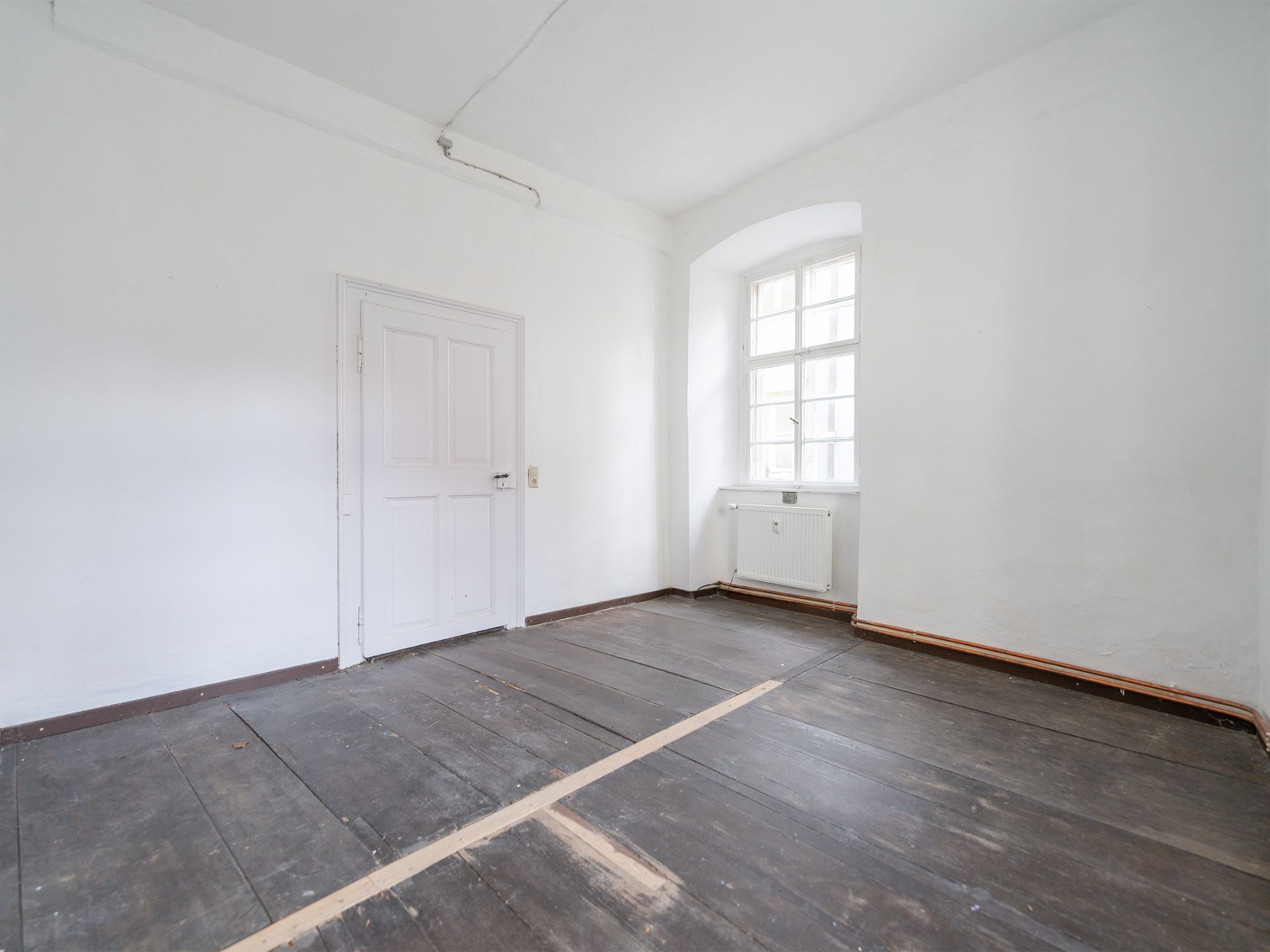 Leerer Raum mit Holzfußboden, weißer Tür und großem Fenster.
