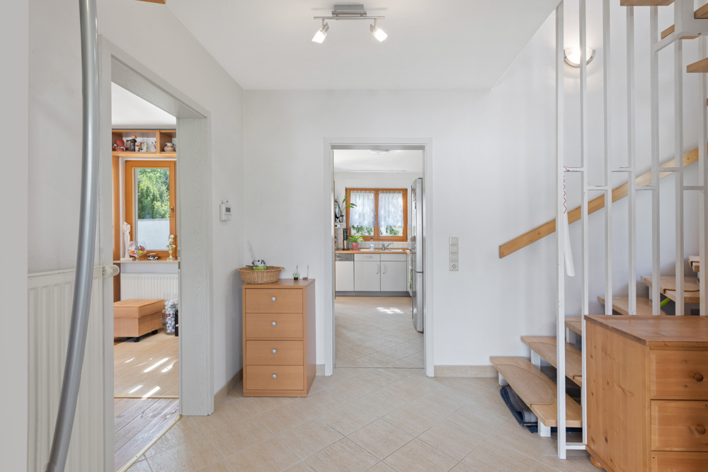 Heller Flur mit Holztreppe, Kommode und Blick in die Küche und ein Wohnzimmer.