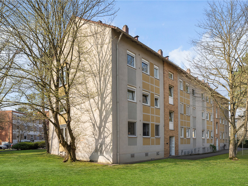 Mehrstöckiges Wohnhaus mit gelber Fassade und mehreren Fenstern, umgeben von Bäumen und Rasen.