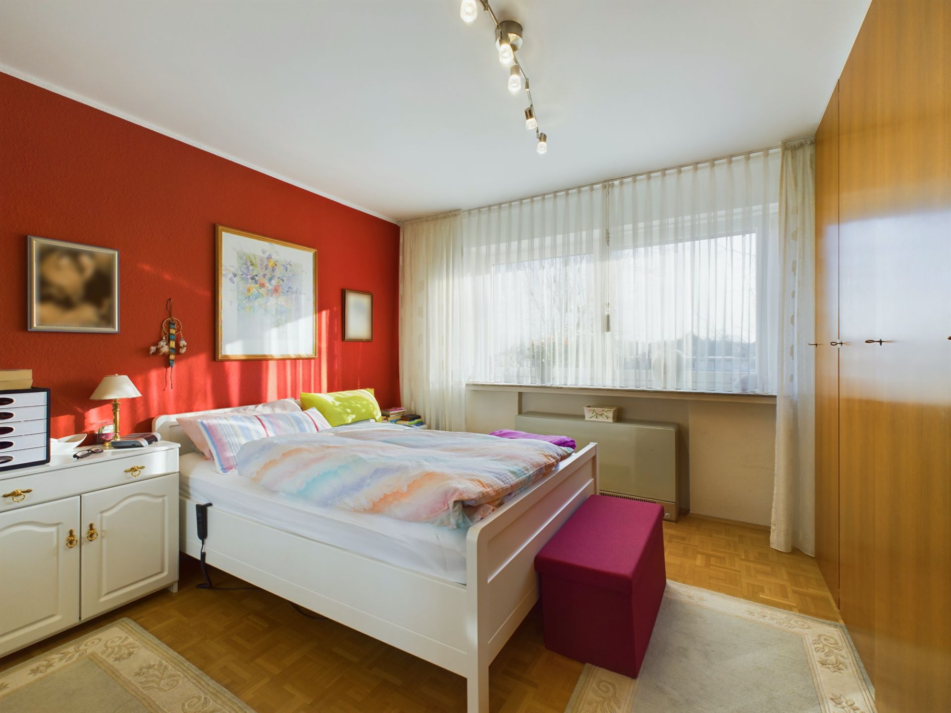 Helles Schlafzimmer mit roter Wand, Doppelbett, weißem Schrank und großem Fenster mit Vorhängen.