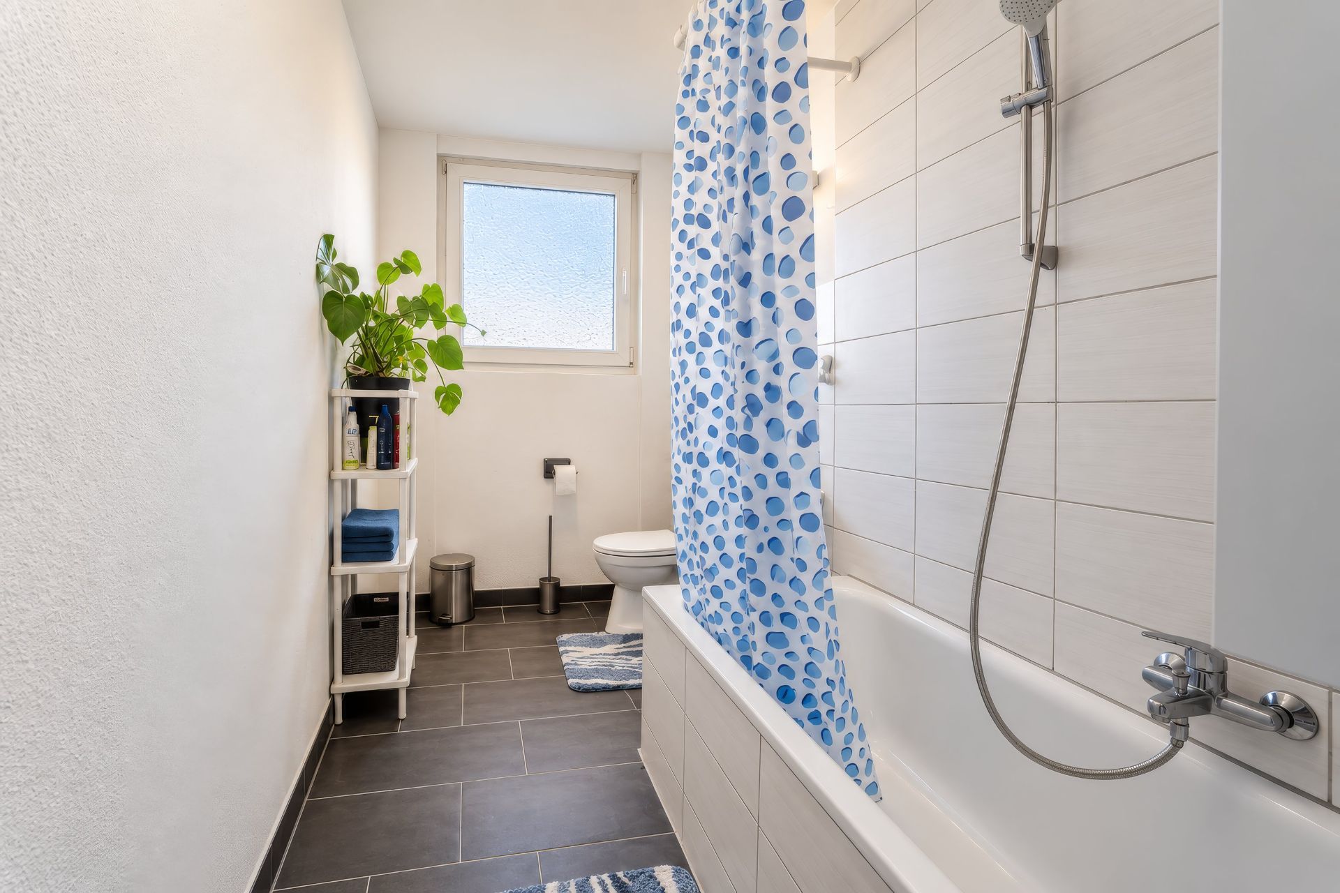 Helles Badezimmer mit Badewanne, Duschvorhang, Regal, Toilette und Fenster.