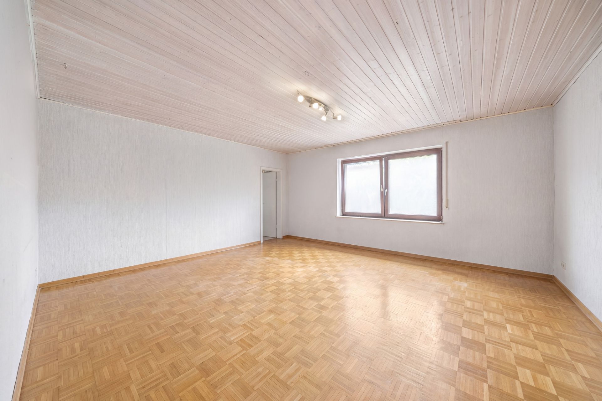 Leerer, heller Raum mit Parkettboden, weißer Holzdecke, Fenster und Tür.