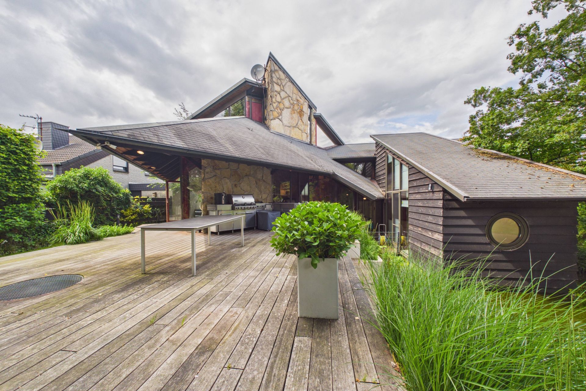 Holzterrasse mit Tisch und Pflanze, modernes Wohnhaus mit Steinfassade und großem Dach.