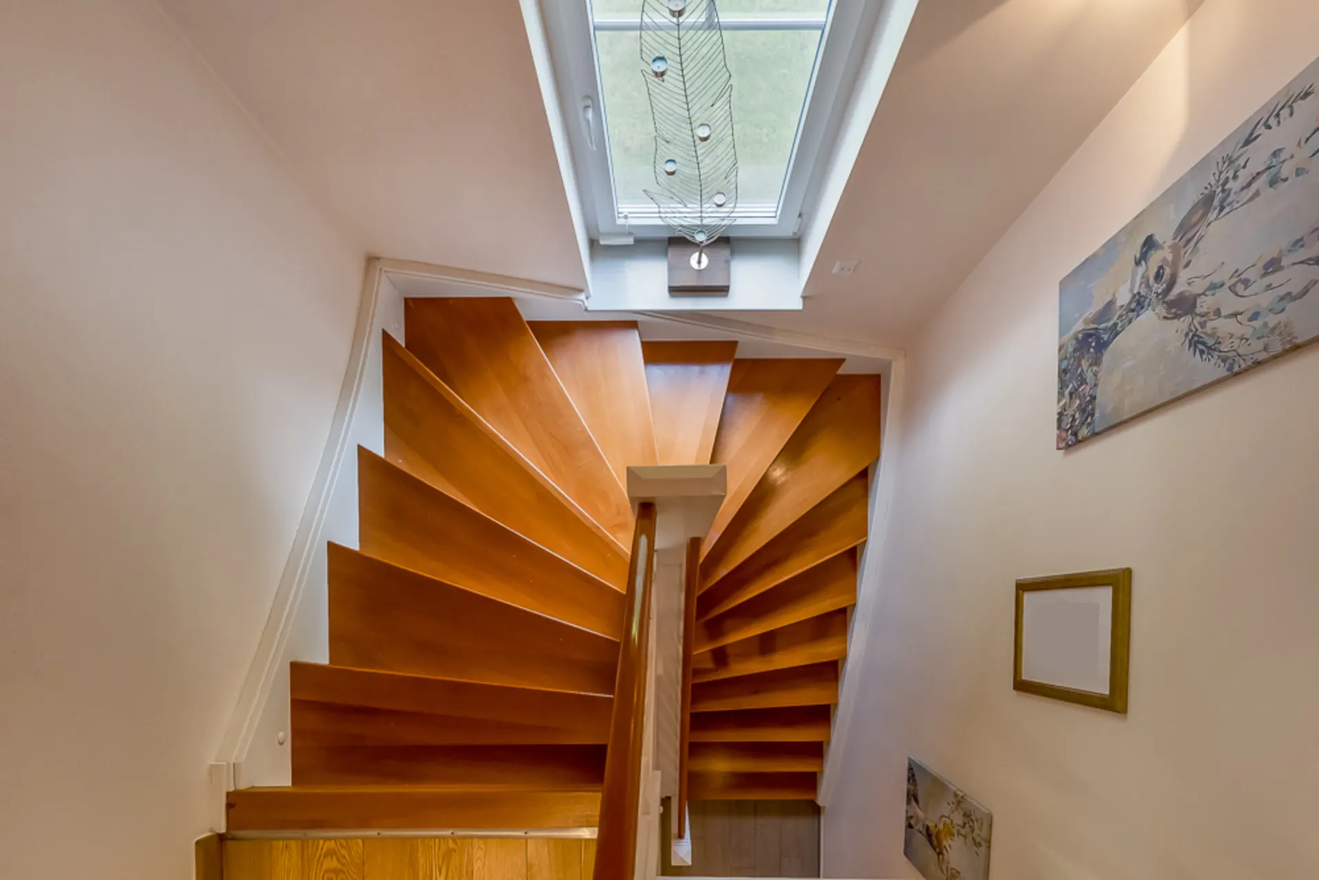 Holztreppe mit Oberlicht und Wandbildern in einem hellen Hausflur.