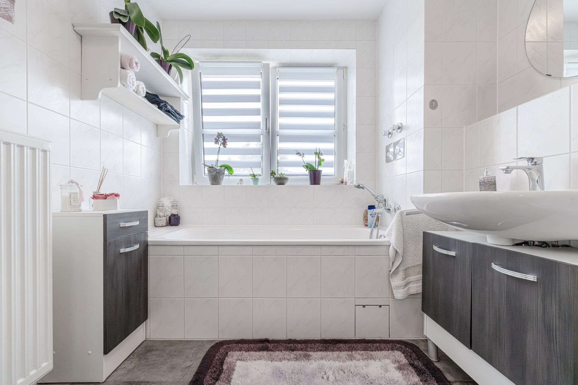 Helles Badezimmer mit Badewanne, Waschbecken, Regal, Pflanzen und zwei Fenstern mit Jalousien.