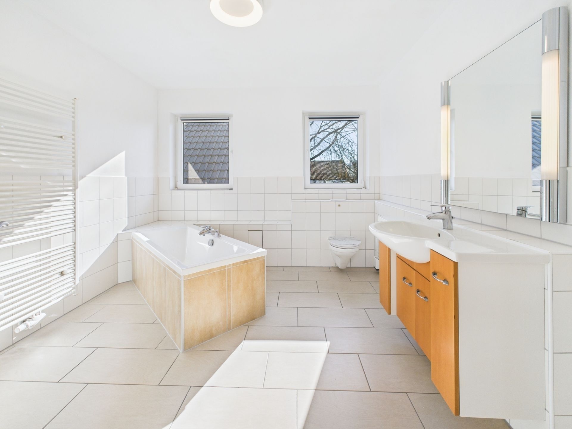 Helles Badezimmer mit Badewanne, Doppelwaschbecken und zwei Fenstern.
