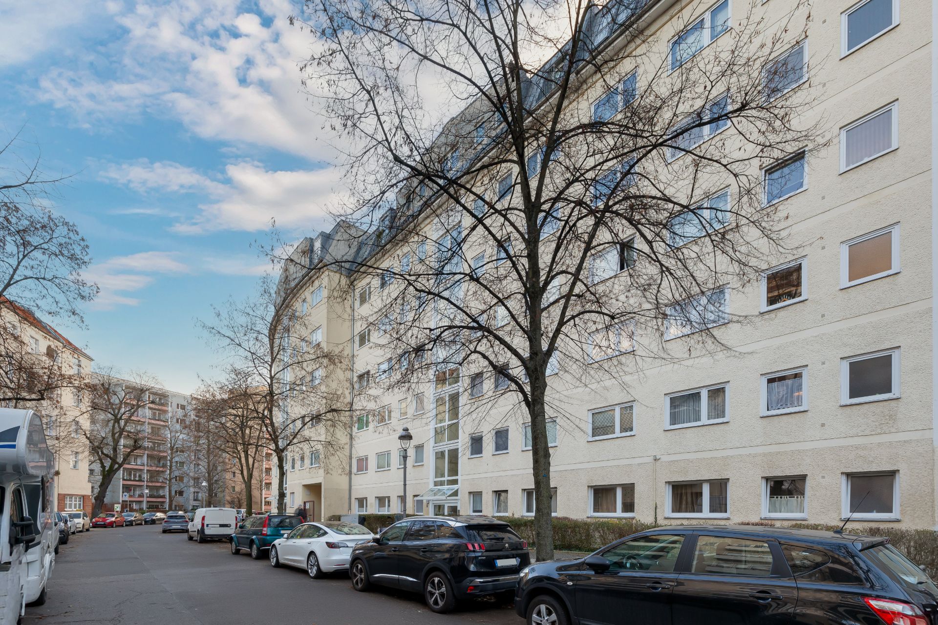 Mehrstöckiges Wohnhaus mit geparkten Autos an einer Straße, kahle Bäume im Vordergrund.