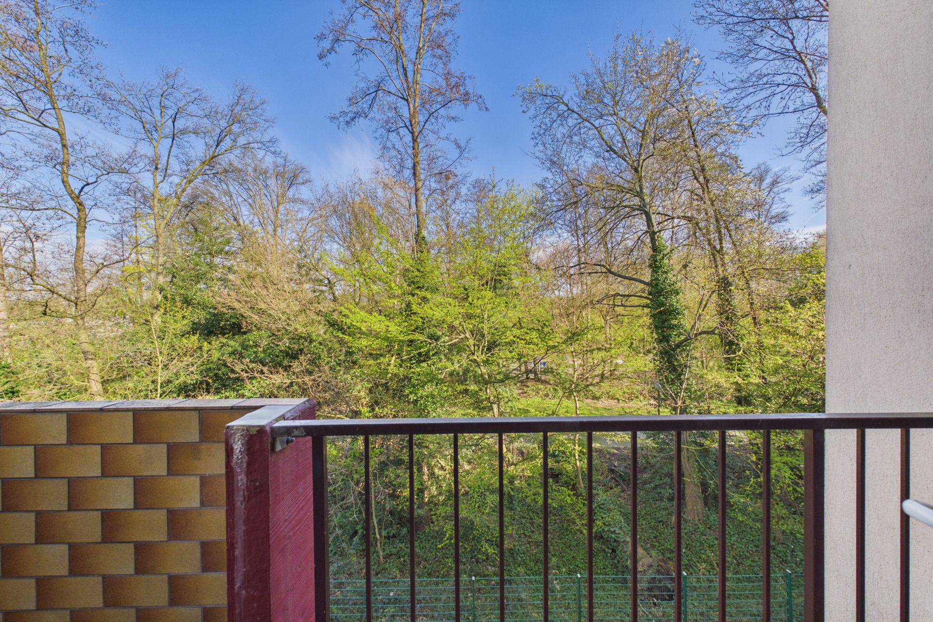 Balkon mit Metallgeländer, Blick auf grüne Bäume und blauen Himmel.