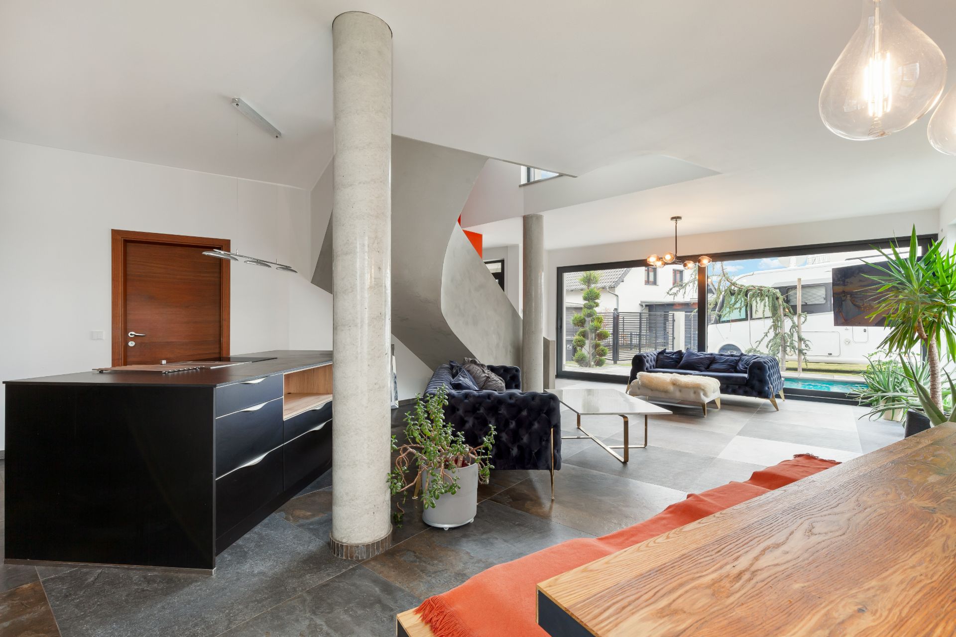 Modernes Wohnzimmer mit offener Küche, großen Fenstern, Pflanzen und einer Wendeltreppe.