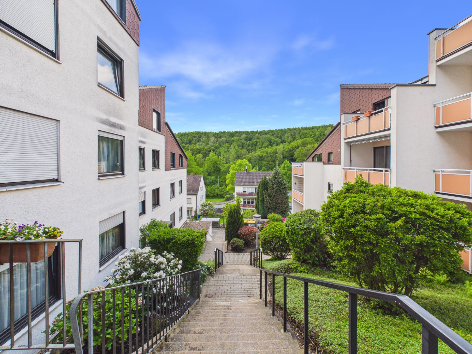 Treppen zwischen Wohngebäuden mit viel Grün und Blick auf einen bewaldeten Hügel.