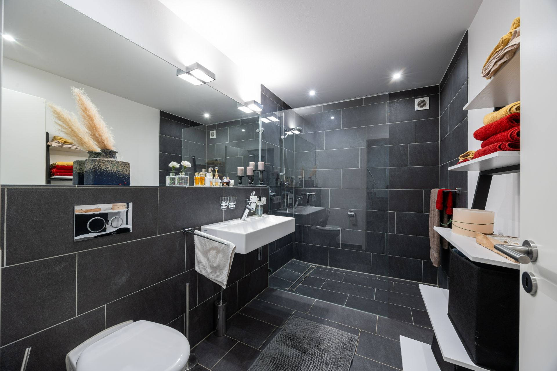 Modernes Badezimmer mit dunklen Fliesen, Waschbecken, Dusche und Regal mit Handtüchern.
