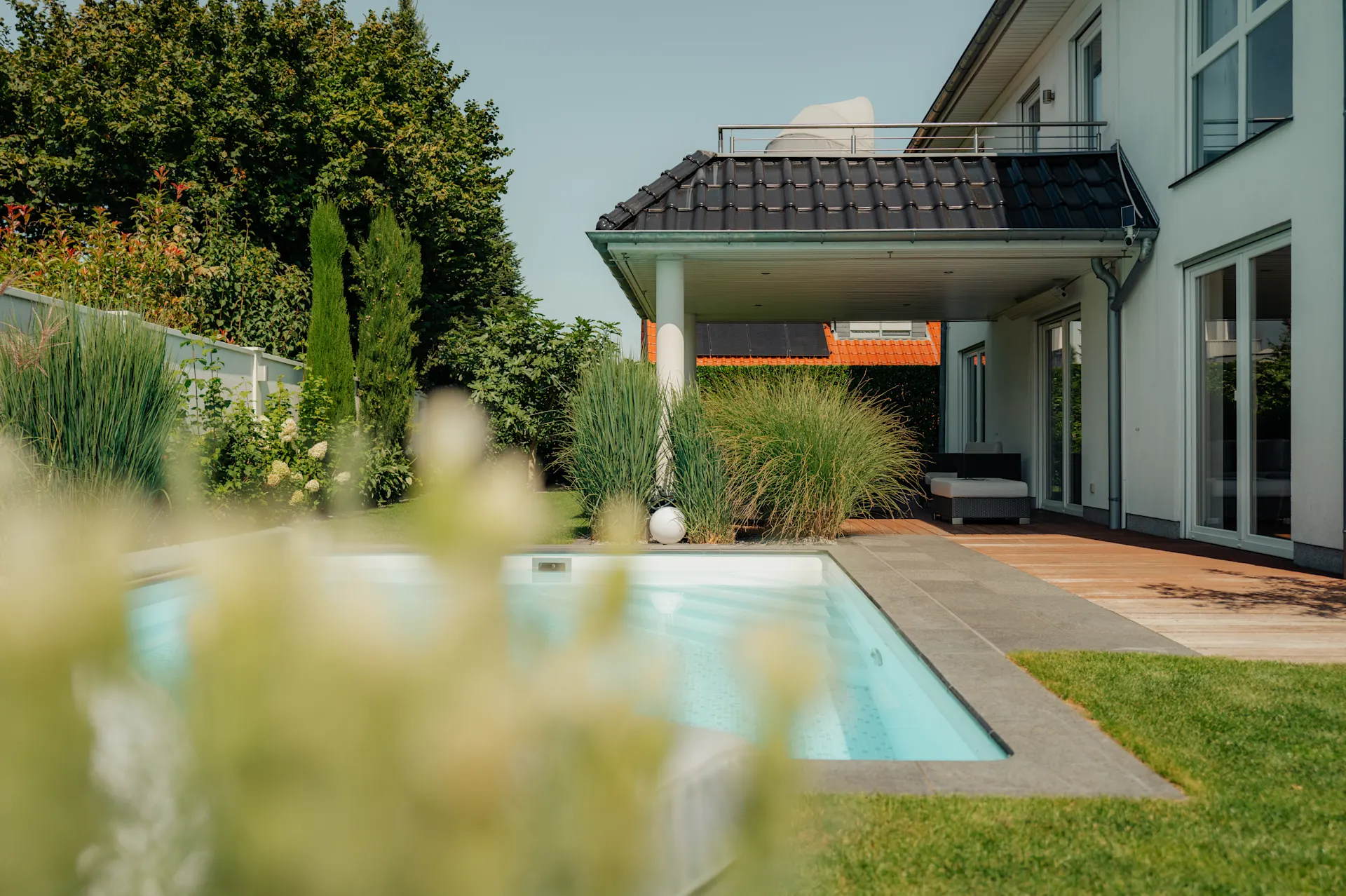 Moderner Garten mit Pool, Terrasse, viel Grün und einem Wohnhaus im Hintergrund.