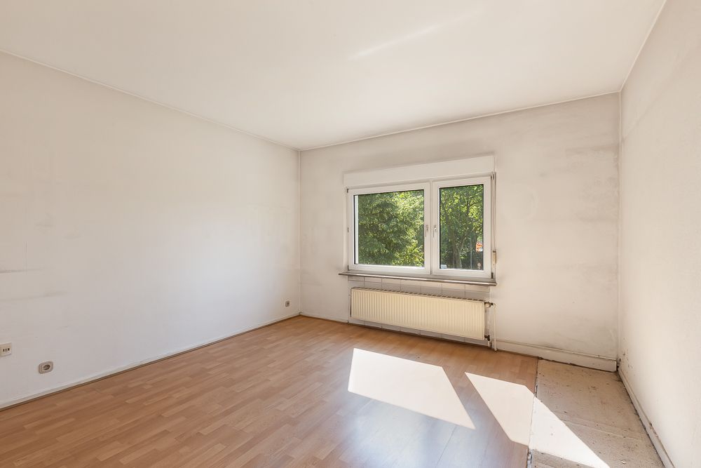 Leeres Zimmer mit großem Fenster, Heizkörper und Laminatboden, Licht fällt auf den Boden.