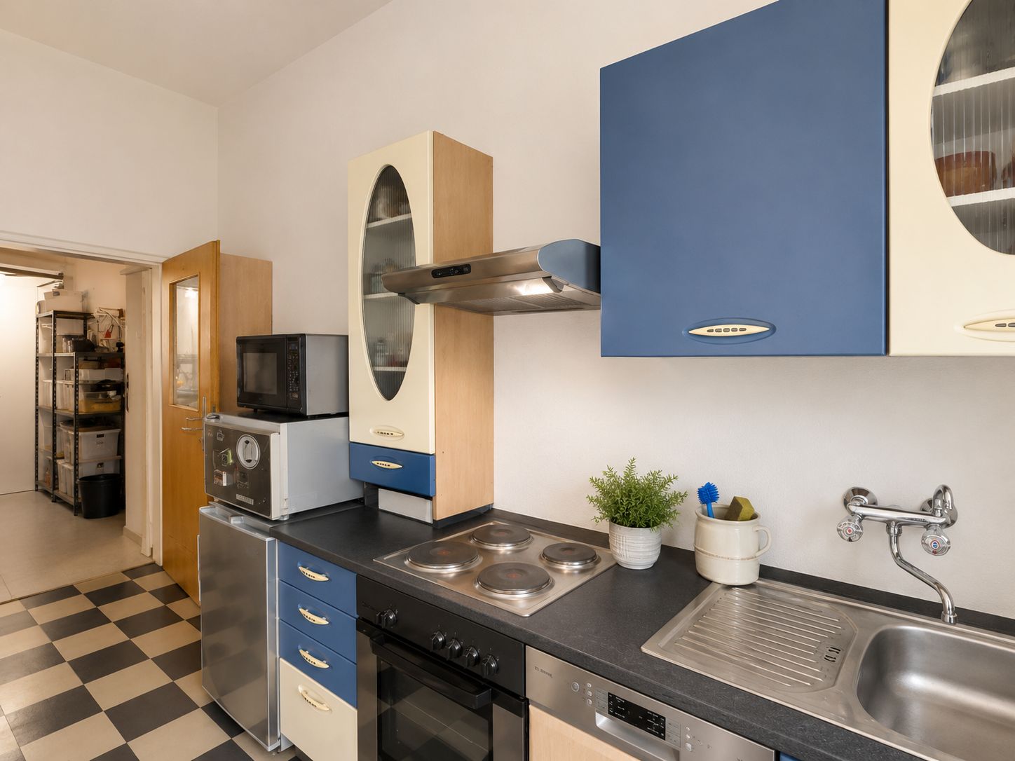 Moderne Küche mit blau-beigen Schränken, Herd, Spüle, Mikrowelle, Pflanze und Küchengeräten.