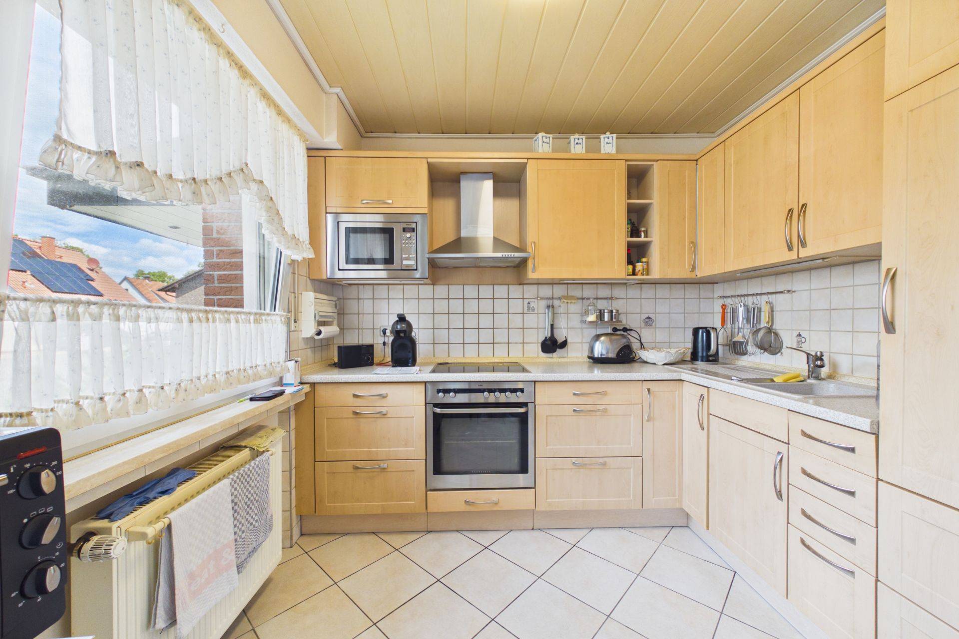 Helle Einbauküche mit Holzschränken, Herd, Spüle und großen Fenstern mit Gardinen.