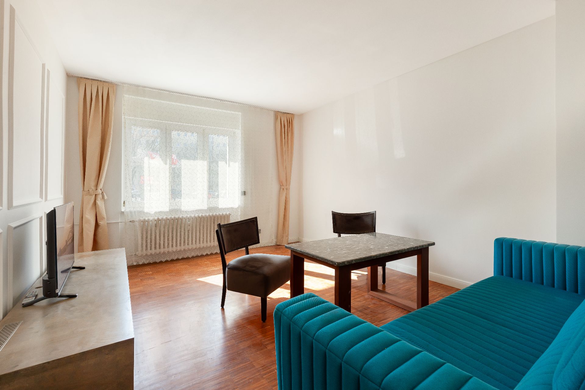 Helles Wohnzimmer mit blauem Sofa, TV, Tisch, zwei Stühlen und großem Fenster mit Vorhängen.