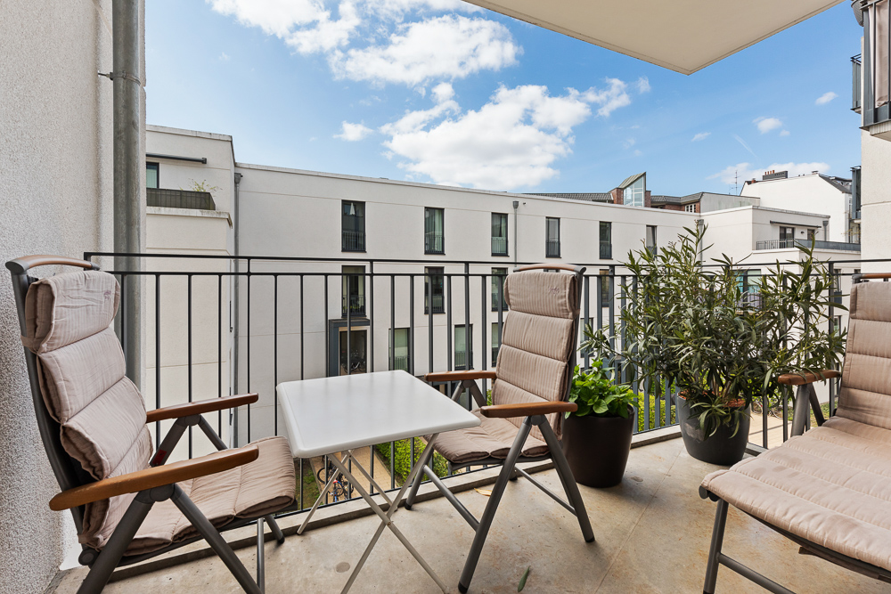Balkon mit drei gepolsterten Stühlen, kleinem Tisch und zwei Pflanzentöpfen, Blick auf modernes Wohngebäude.