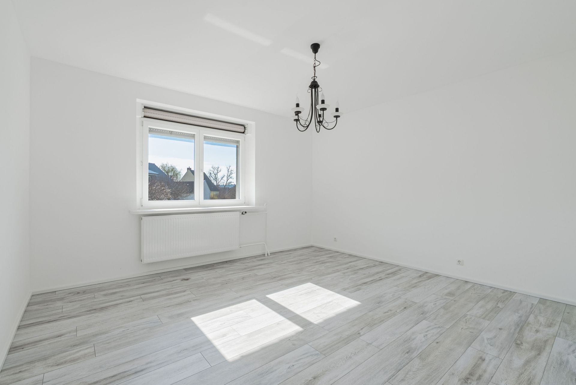 Helles, leeres Zimmer mit Fenster, weißem Boden und schwarzem Kronleuchter.