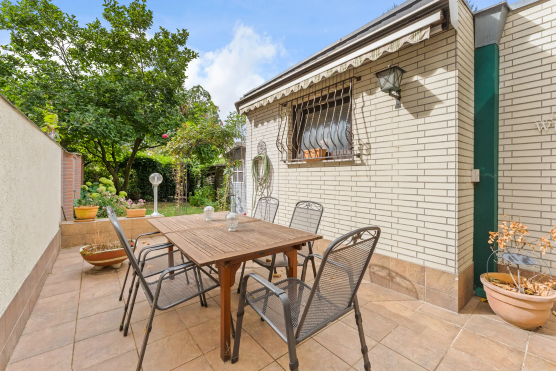 Terrasse mit Holztisch, sechs Stühlen und Pflanzen, daneben ein Garten mit Bäumen.