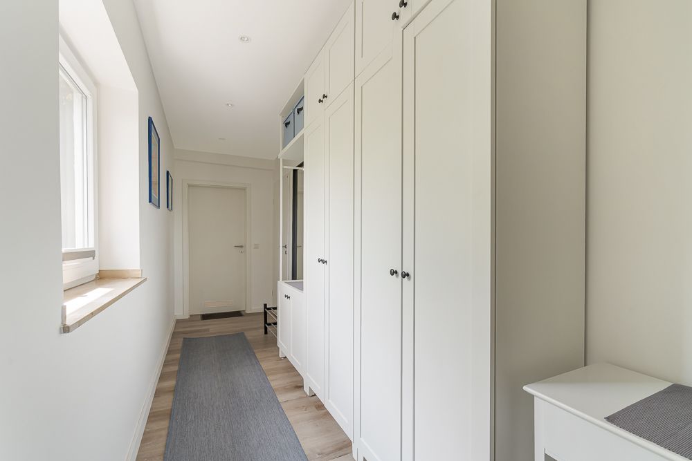 Heller Flur mit weißen Einbauschränken, Fenster links und Teppichläufer auf Holzfußboden.