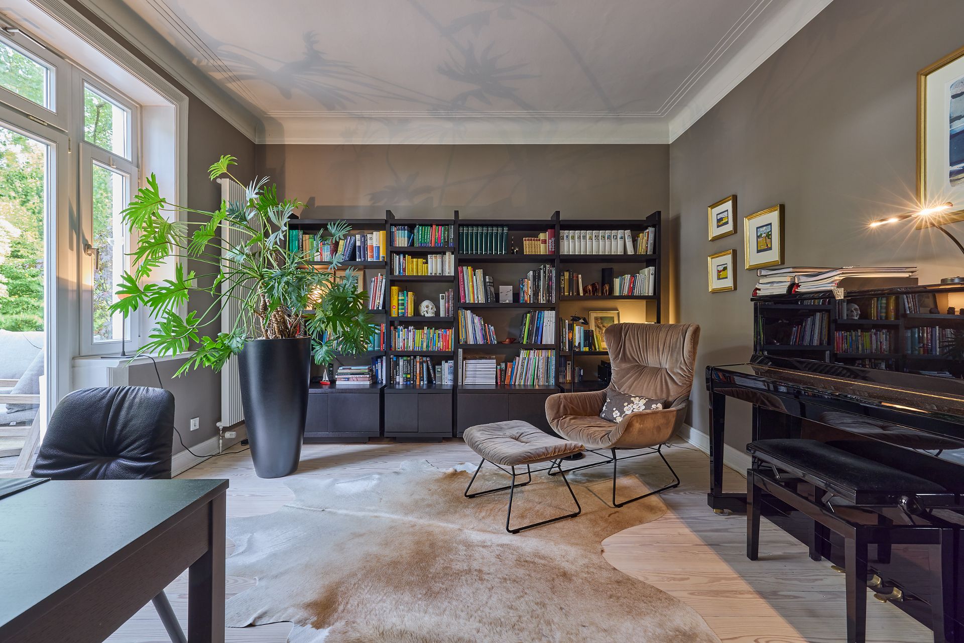 Helles Wohnzimmer mit Bücherregal, Sessel, Klavier, großem Pflanzentopf und Teppich.